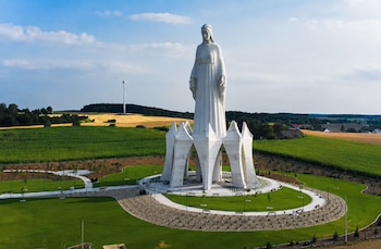 Estatua blanca de figura femenina sobre pedestal con arcos. Césped, caminos, árboles, campos cultivados y aerogenerador bajo cielo azul con nubes blancas