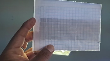Una mano sostiene ante la luz una hoja cuadriculada sellada con cinta transparente que contiene pequeños objetos oscuros y rectangulares