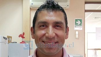 Primer plano de un hombre sonriente con cabello oscuro, camisa de cuello, en un interior con un pequeño ventilador rojo y una señal de salida en segundo plano