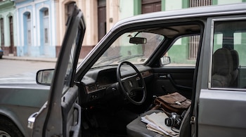 Interior de un auto gris con la puerta abierta, un bolso de cuero, papeles y una cámara sobre el asiento frente a edificios coloniales.