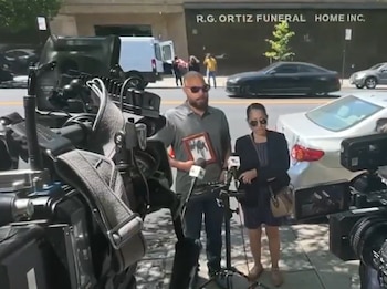 Un hombre con un cuadro y una mujer permanecen frente a micrófonos y cámaras de televisión en una acera cerca de varios automóviles