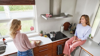 Una mujer corta vegetales junto a la ventana de la cocina mientras una joven está sentada sobre la encimera
