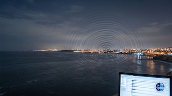 Vista nocturna de una costa con luces urbanas, ondas concéntricas sobre el agua y una pantalla con gráficos de ondas y un logotipo circular.
