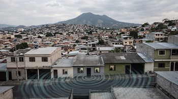 Casas de baja altura en una calle urbana con ondas concéntricas en el asfalto y una montaña al fondo bajo un cielo nublado.
