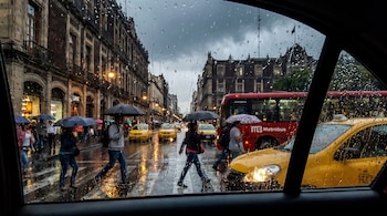 Vista a través de una ventana mojada por la lluvia; en la calle se observan edificios, farolas encendidas, personas con paraguas, taxis amarillos y un Metrobús rojo.