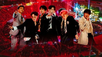 Los miembros del grupo BTS con trajes formales frente a la imagen de un estadio iluminado de rojo con multitud de asistentes.