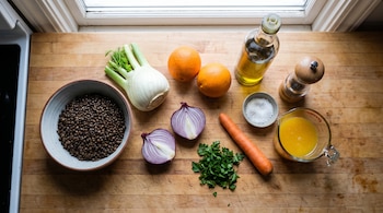 Un tazón con lentejas, hinojo, naranjas, cebolla morada, zanahoria, perejil, aceite de oliva, sal y jugo sobre una mesada de madera.