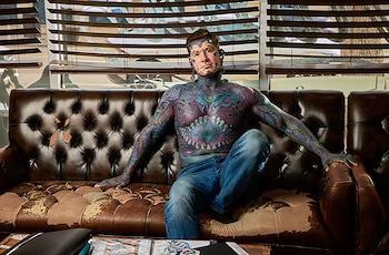 Un hombre sin camiseta con tatuajes en el cuerpo y rostro sentado en un sofá de cuero gastado frente a una ventana con persianas