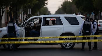 Camioneta SUV blanca con puerta abierta, cristal trasero lateral roto. Cinta amarilla "PROHIBIDO EL PASO". Dos hombres con chalecos. Tres siluetas en el interior, una mano con pistola.