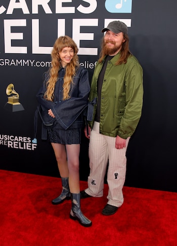 Los músicos Livvy Bennett y Michael B. Hunter. La mujer llevó un conjunto en tonos oscuros compuesto por una chaqueta amplia, un vestido corto y botas de media caña con un diseño brillante. Su cabello largo y ondulado cayó suelto sobre sus hombros, y su sonrisa le dio un toque relajado a su apariencia. El hombre, a su lado, vistió una chaqueta verde con una camisa oscura, pantalones beige con detalles gráficos y zapatos negros. Completó su look con un gorro oscuro y una barba que destacó como elemento característico