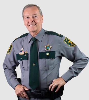 Hombre de mediana edad con uniforme gris de sheriff, camisa, corbata verde oscura, insignias, con las manos en la cintura y sonriendo