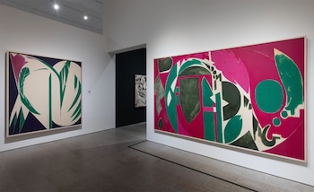 Dos grandes pinturas abstractas de colores verde, rosa y morado cuelgan en las paredes blancas de una galería iluminada