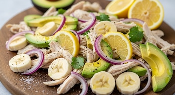 Primer plano de una ensalada de pollo desmenuzado, rodajas de banana, palta, cebolla morada y limón, espolvoreada con pimienta y sal, sobre una tabla de madera.