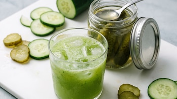Vaso de jugo de pepino con hielo, frasco abierto de pepinillos, rodajas de pepino fresco y encurtido sobre una superficie blanca.