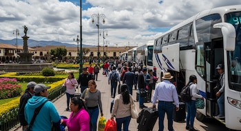 Vista de una multitud abordando varios autobuses blancos de pasajeros estacionados en una plaza urbana con jardines, una fuente y edificios históricos al fondo.