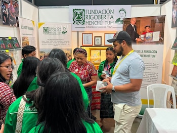 Rigoberta Menchú Tum rodeada de personas en un stand con banners de su fundación y estantes con libros. Un hombre sostiene un libro