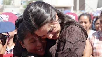 Primer plano de Andrea Chávez, con blusa marrón bordada, sonriendo mientras abraza a una mujer de tez morena que también sonríe con los ojos cerrados. Fondo con gente