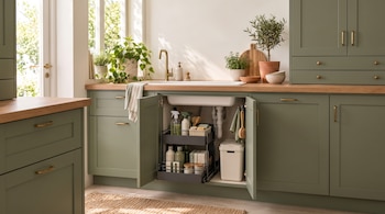 Armario de cocina verde con puertas abiertas que contiene un organizador metálico con productos de limpieza y un cubo de basura.