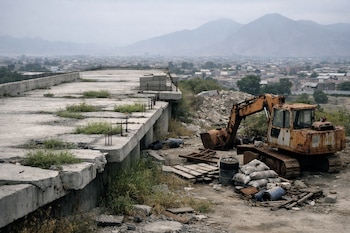 Un terraplén de concreto inacabado con vegetación y barras de refuerzo se extiende junto a una excavadora oxidada, con casas y montañas al fondo bajo un cielo nublado.