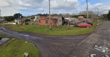 Vista de una esquina con calles de tierra y pasto, casas de ladrillo y techos de chapa, postes de luz, un auto rojo y otro oscuro, y cielo nublado