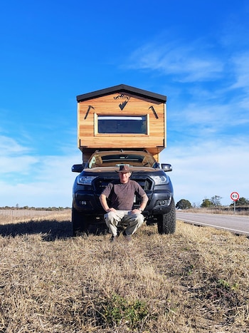 Hombre en cuclillas frente a una camioneta pickup negra con un módulo de cámper de madera. Al fondo hay un campo seco y una carretera con señal de 60