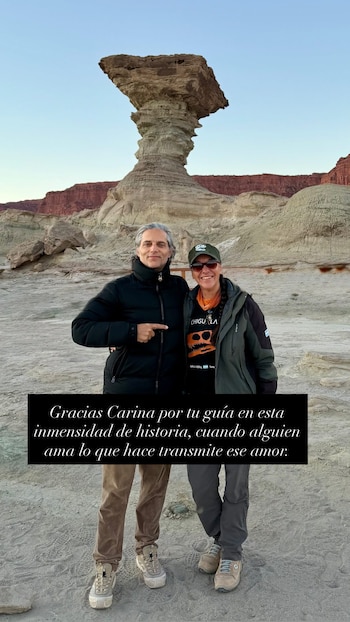 Dos personas de pie, un hombre y una mujer, frente a una formación rocosa alta en un paisaje árido con montañas y cielo despejado. Texto superpuesto