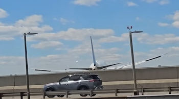 La cola y alas de un avión de United Airlines se ven detrás de una barrera de hormigón; un SUV plateado transita en primer plano por una autopista bajo cielo azul