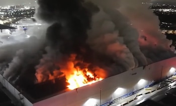 Vista aérea nocturna de un edificio industrial con grandes llamas y denso humo que se eleva desde el techo. Luces de la ciudad en el fondo