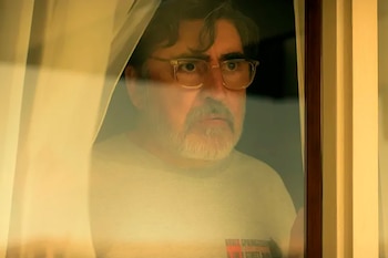 Alfred Molina, hombre de mediana edad con barba canosa y gafas claras, mira con expresión seria a través de una ventana empañada