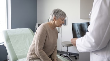 Mujer adulta sentada en camilla de consultorio médico observa radiografía de huesos que sostiene un médico con bata blanca.