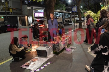 Personas junto a una figura de cartón, velas, flores, cartas y fotografías en una acera nocturna con un autobús al fondo