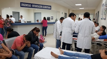 Vista de una sala de urgencias de hospital con letrero 'Intoxicación Alimentaria', donde pacientes esperan y médicos de bata blanca asisten a otros en camillas.