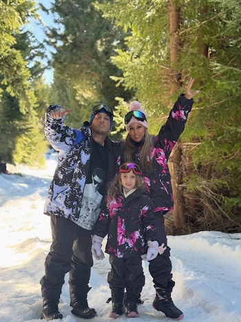 Un hombre, una mujer y una niña con ropa de nieve y antiparras posan en un camino cubierto de nieve rodeado de árboles