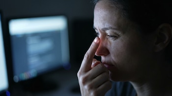 Primer plano del rostro de una mujer en oscuridad, con el dedo índice derecho cerca de su ojo, y un monitor de computadora borroso detrás.