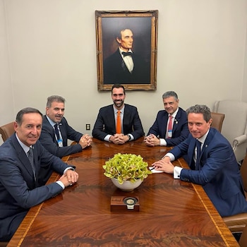 Cinco hombres en traje sentados alrededor de una mesa de madera con un centro de flores verdes, bajo un retrato de Abraham Lincoln