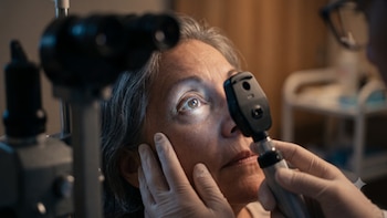 Médico con guantes blancos examina el ojo de una paciente con un oftalmoscopio iluminado. Equipo de examen ocular en primer plano y de fondo borroso