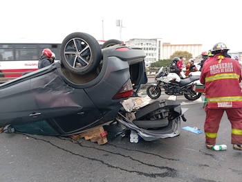Coche oscuro volcado de lado en una carretera, con un neumático visible y partes del maletero abiertas. Bomberos y una motocicleta de emergencia están cerca