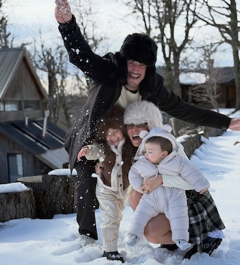 Una familia sonriente de cuatro, dos adultos y dos niños, vestidos de invierno, juegan en la nieve con cabañas de madera al fondo