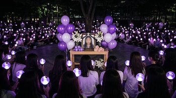 Gran grupo de personas sentadas en un círculo por la noche, sosteniendo army bombs encendidas de luz morada alrededor de un altar con globos, flores y una foto borrosa.