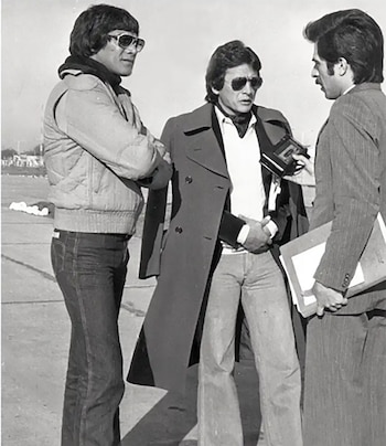 Una fotografía en blanco y negro muestra a tres hombres al aire libre. Dos hombres con gafas de sol, uno con chaqueta acolchada y otro con gabardina, conversan con un tercero que sostiene una grabadora