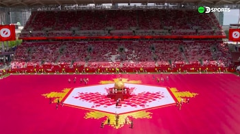 Vista aérea de un estadio lleno con un campo rojo vibrante, adornado con un diseño de hoja de arce blanco y dorado, una esfera central y artistas