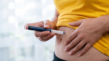 Primer plano de una mano sujetando un bolígrafo de inyección azul insertado en el abdomen de una mujer con camiseta amarilla.