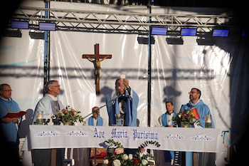 Un sacerdote eleva una hostia ante un altar con un crucifijo, arreglos florales y la frase Ave Maria Purisima junto a otros religiosos