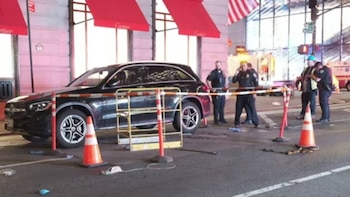 Un SUV compacto oscuro estacionado en la calle con conos naranjas y barreras. Oficiales de policía y una ambulancia son visibles al fondo