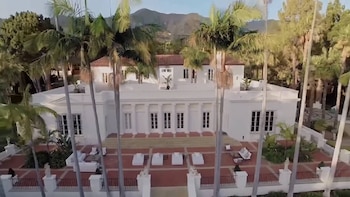 Vista aérea de una gran mansión blanca con tejado rojo, múltiples ventanas y un patio. Palmeras altas en primer plano y alrededor, montañas al fondo