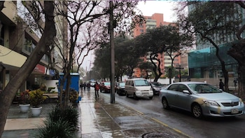 Una calle con la pista mojada, vehículos en fila, peatones en la acera, árboles a los lados y edificios bajo un cielo gris