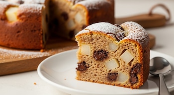 Primer plano de una rebanada de budín de pera y chocolate en un plato blanco, con el resto del budín en el fondo. Está espolvoreado con azúcar.
