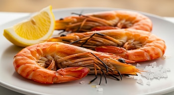 Tres gambas enteras asadas con cabeza y cáscara se presentan en un plato blanco con sal gruesa y un trozo de limón, destacando su color naranja.