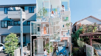 Un edificio de vidrio de varios niveles con tres personas, plantas en macetas y un automóvil azul estacionado, flanqueado por otras casas bajo un cielo despejado.