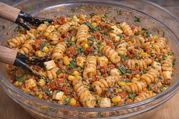 Tazón de vidrio con ensalada de pasta, salsa rojiza, granos de maíz, cubos de queso, tomate seco y perejil. Dos utensilios de madera para servir.